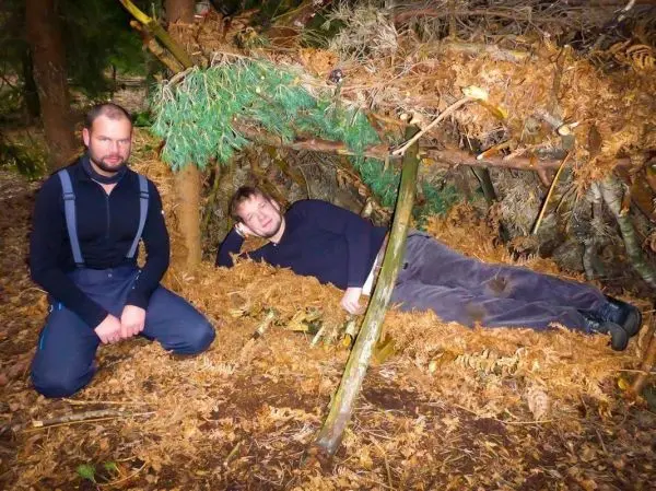 Zwei Personen neben einer selbstgebauten Tarp-Notunterkunft aus Ästen und Blättern
