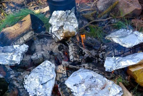 Alufolie-Päckchen liegen auf glühenden Kohlen neben einem Kessel über dem Feuer