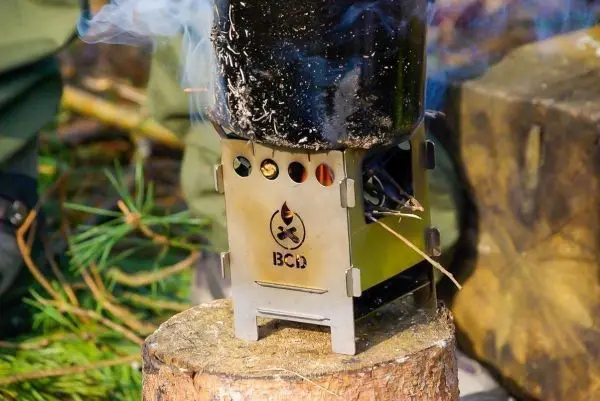 Hobo-Kocher auf Baumstamm mit rauchendem Kessel und brennendem Zunder