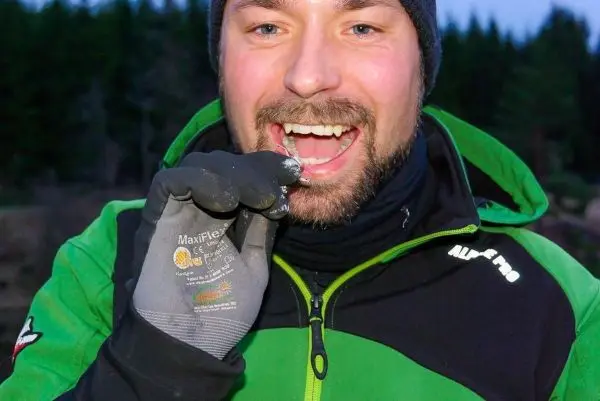 Mann hält etwas zwischen den Zähnen und lächelt, trägt Handschuhe und eine Jacke