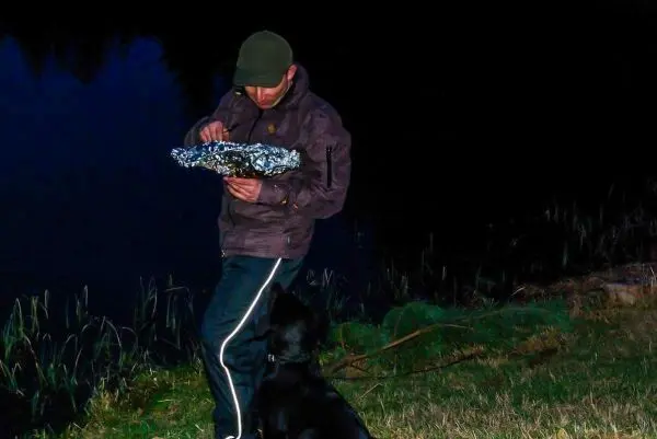Mann hält mit beiden Händen ein in Alufolie gewickeltes Objekt am Ufer eines Gewässers