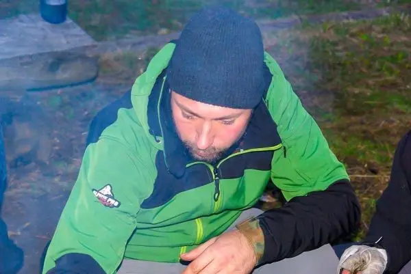 Mann sitzt am Boden und hantiert mit Zunder in der Nähe eines kleinen Feuers