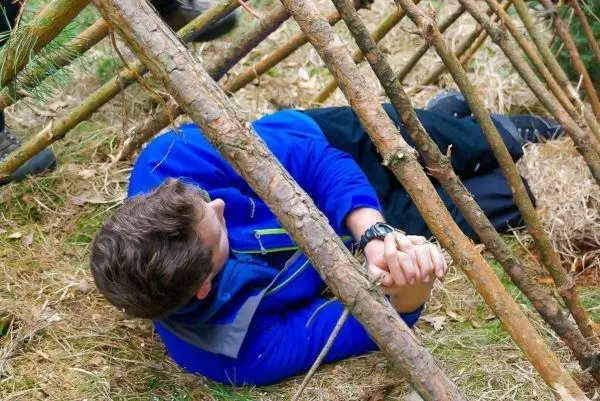 Person liegt unter einem selbstgebauten Unterstand aus Ästen