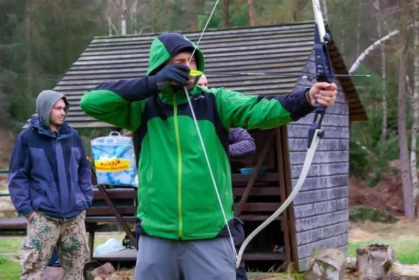 Person zielt mit einem Bogen auf ein Ziel, im Hintergrund eine Holzhütte