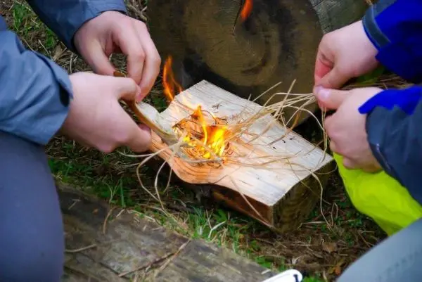 Zunder glimmt auf einem Holzstück, während zwei Hände ihn bearbeiten