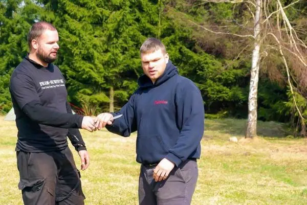 Zwei Personen führen eine Übergabe eines Messers in einer Waldumgebung durch