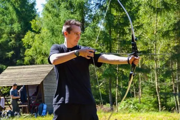 Bogen wird gespannt, während im Hintergrund eine Holzhütte sichtbar ist
