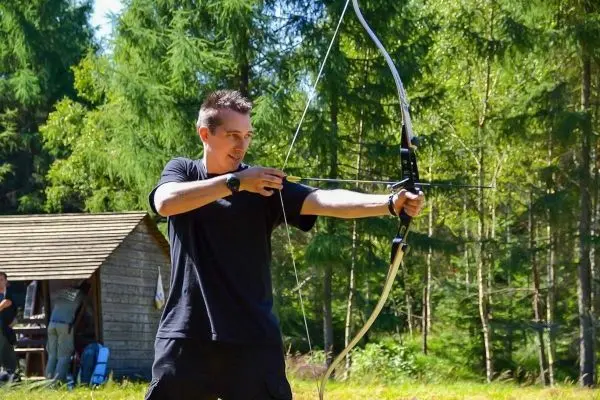 Bogen wird gespannt, während im Hintergrund eine Holzhütte sichtbar ist