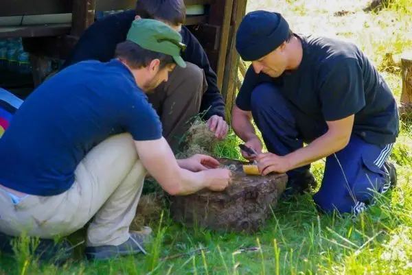 Drei Personen bearbeiten einen Holzblock mit Werkzeugen beim Überlebenstraining