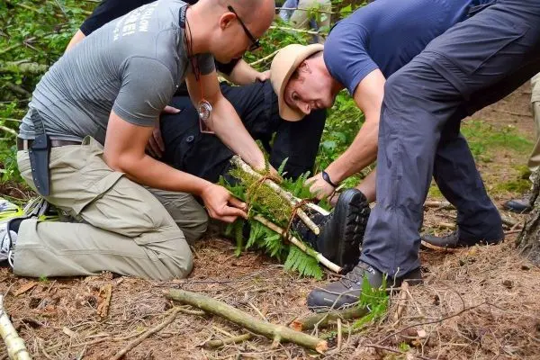 Drei Personen stabilisieren ein Bein mit improvisierter Schiene aus Ästen und Farn