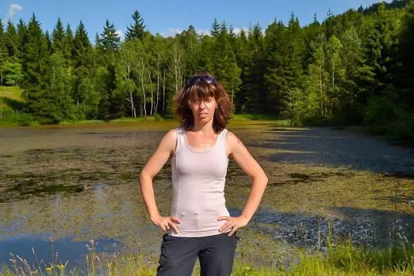 Frau steht mit Händen in der Hüfte am Ufer eines Gewässers, umgeben von Bäumen