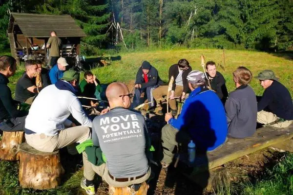 Gruppe sitzt um ein kleines Feuer auf Baumstümpfen und hält Stockstücke