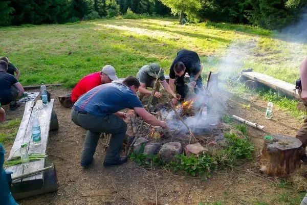 Gruppenmitglieder fügen Holz zu einem kleinen Bodenfeuer hinzu, Rauch steigt auf