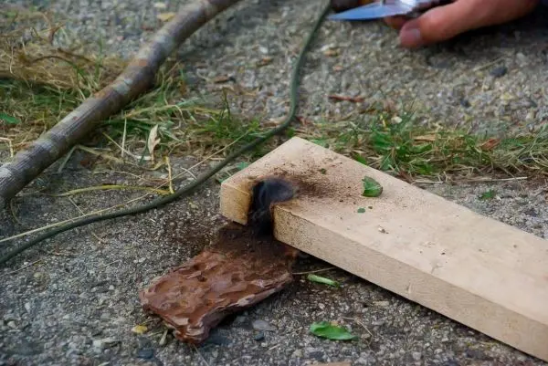 Holzstück mit Kerbe und glühender Stelle, daneben Zunder und Werkzeug sichtbar