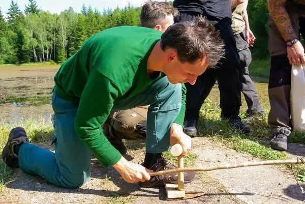 Mann bearbeitet Holzstück mit einem Stock und einem runden Gegenstand auf dem Boden