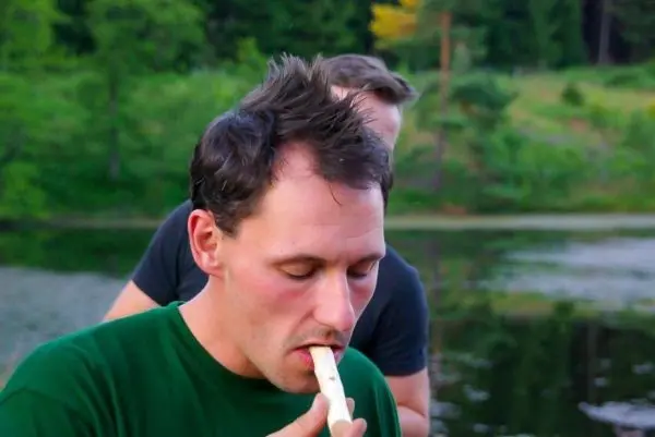 Mann bläst in ein selbstgebautes Blasinstrument am Ufer eines Gewässers