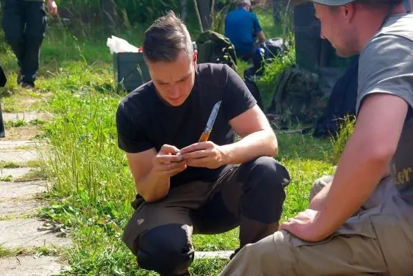 Mann sitzt im Gras und schnitzt mit einem Messer an einem Holzstück