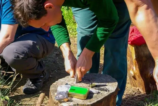 Messer schneidet Holzstück auf einem Baumstumpf, Ausrüstung daneben sichtbar