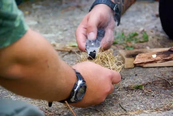 Person hält ein Messer über trockenes Gras beim Feuer machen