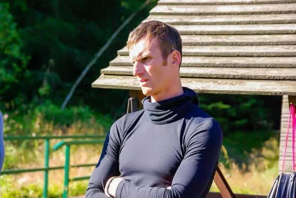 Person steht mit verschraenkten armen neben einem holzgebaeude