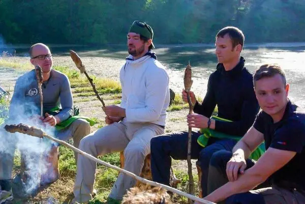 Vier Personen sitzen am Ufer, halten Stockspieße über einem kleinen Feuer