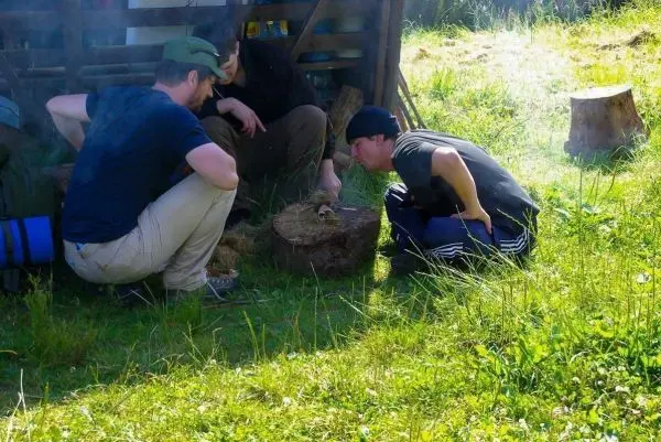 Zunder glimmt auf einem Holzstück, während zwei Personen konzentriert darüber beugen