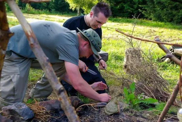 Zwei Personen arbeiten am Boden, einer schnitzt mit einem Messer, der andere beobachtet