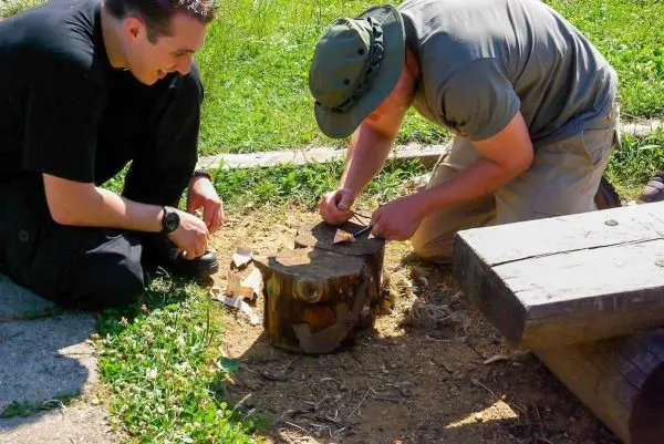 Zwei Personen bearbeiten einen Holzstamm mit einem Messer zur Zunderherstellung