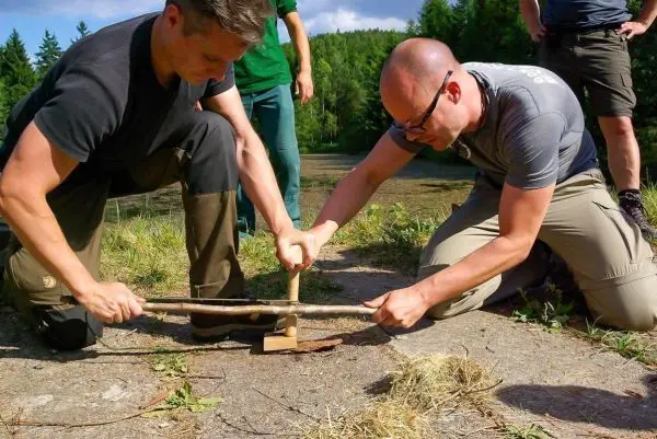 Zwei Personen nutzen eine Feuerbohrmaschine, um Zunder zu entzünden
