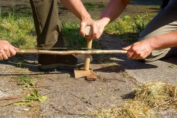 Zwei Personen nutzen einen Feuerbohrer zur Zundererzeugung auf dem Boden