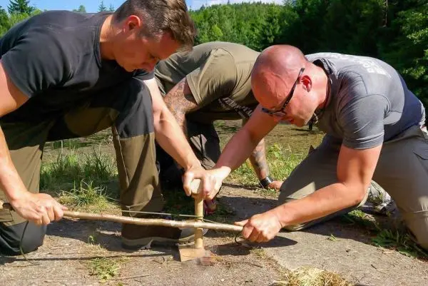Zwei Personen nutzen einen Feuerbohrer zur Zundererzeugung auf dem Boden