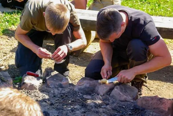 Zwei Personen schnitzen mit Messern an einem Feuerplatz aus Steinen