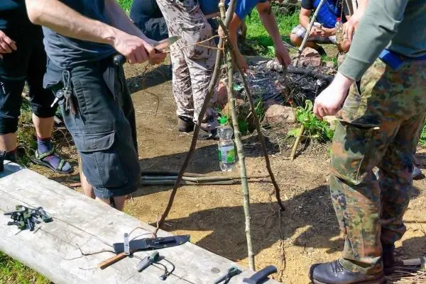 Drei Personen bauen ein Dreibein aus Ästen über einem Feuerplatz