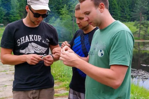 Drei Personen bereiten Zunder für ein Feuer am Ufer eines Gewässers vor