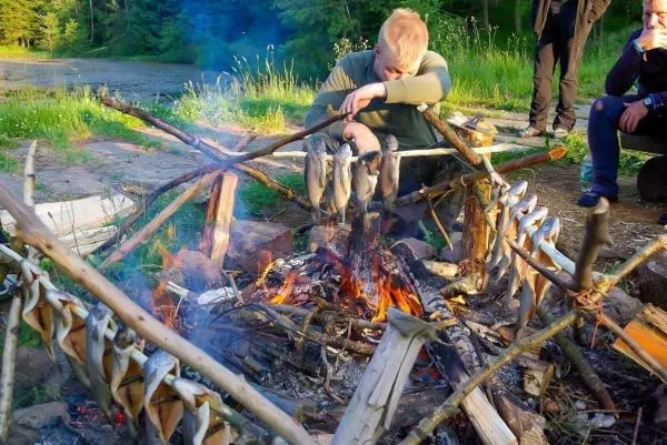 Fische hängen über einem offenen Feuer zum Garen, während Personen in der Nähe sitzen