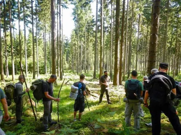 Gruppe mit Rucksäcken steht im Wald und hört einem Anleiter zu