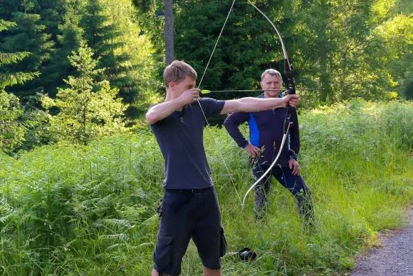 Junge spannt einen Bogen im Wald, ein weiterer Mann beobachtet