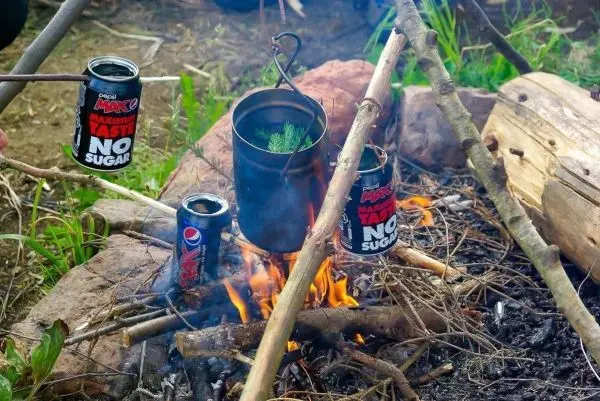 Kessel hängt über Feuerstelle, zwei Dosen auf Ästen neben dem Feuer