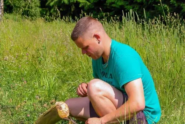 Mann bearbeitet Holzstück mit einem Messer im hohen Gras