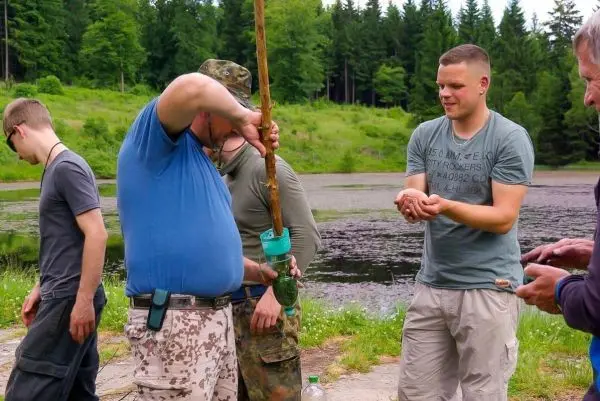 Mann mit Holzstock und Wasserbehälter steht am Ufer eines Gewässers