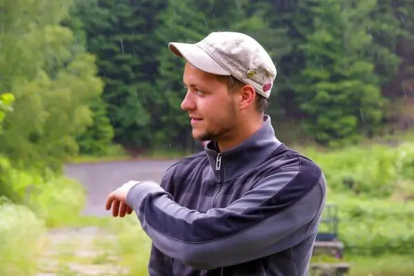 Mann mit Kappe und Jacke steht im Freien, umgeben von Bäumen