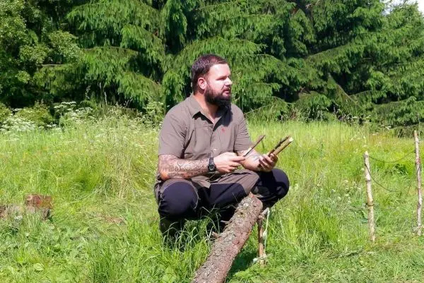 Mann sitzt im Gras und bearbeitet Holzstücke mit einem Messer