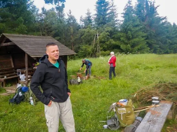 Mann steht neben Rucksack, während andere Holzstücke bearbeiten und eine Hütte im Hintergrund sichtbar ist