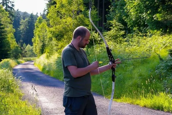 Person spannt einen Bogen auf einem Weg zwischen Bäumen