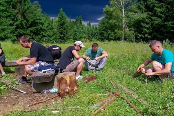 Teilnehmer bearbeiten Holzstücke mit Messern auf Baumstämmen im Freien