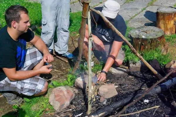 Zunder glimmt in einem kleinen Bodenfeuer, während zwei Personen daneben arbeiten