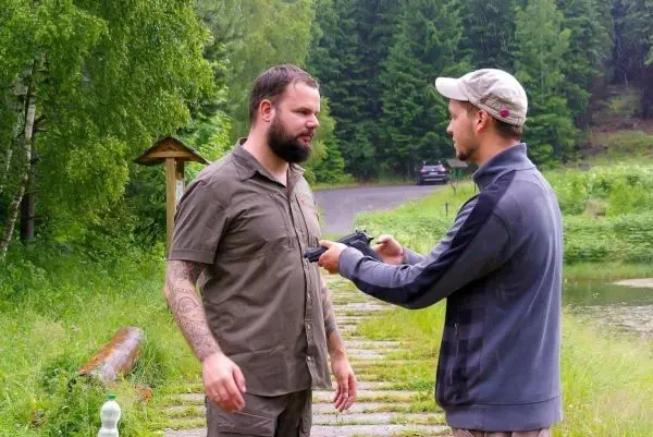 Zwei Männer stehen an einem Weg, einer übergibt eine Handfeuerwaffe
