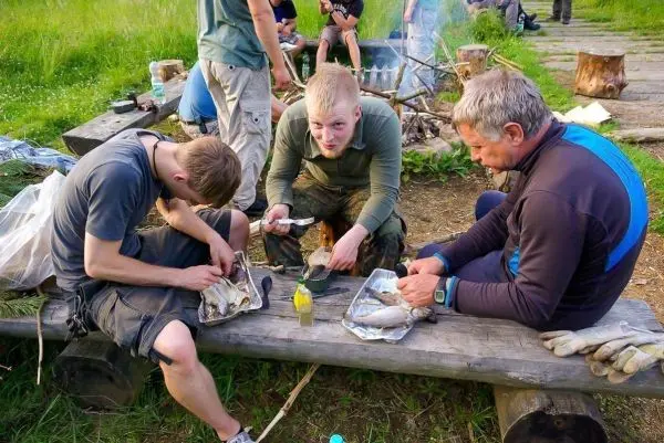 Zwei Personen filetieren Fische auf einer Holzbank, während im Hintergrund ein Feuer brennt