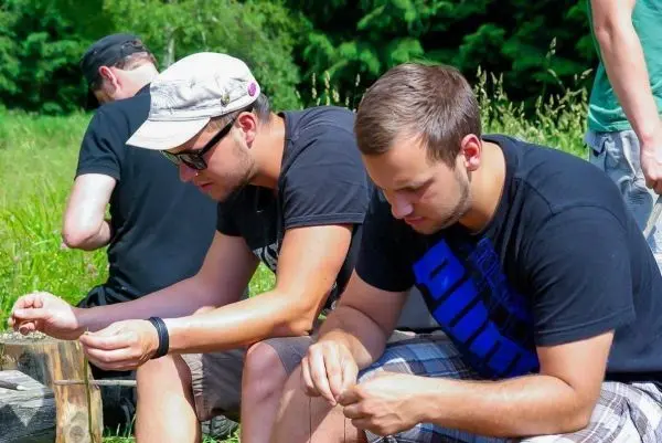 Zwei Personen knoten Schnüre, während sie auf einem Holzstück sitzen
