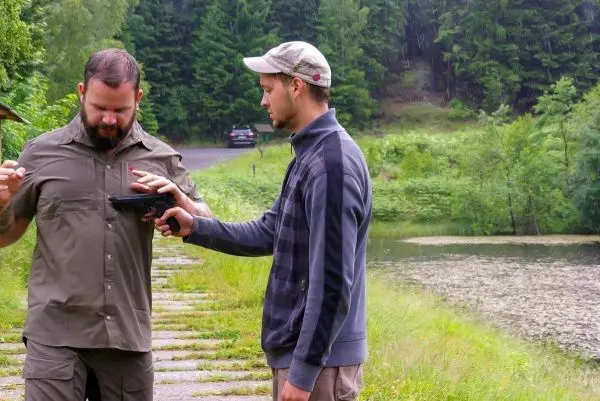 Zwei Personen stehen an einem Gewässer, eine hält eine Pistole auf die andere